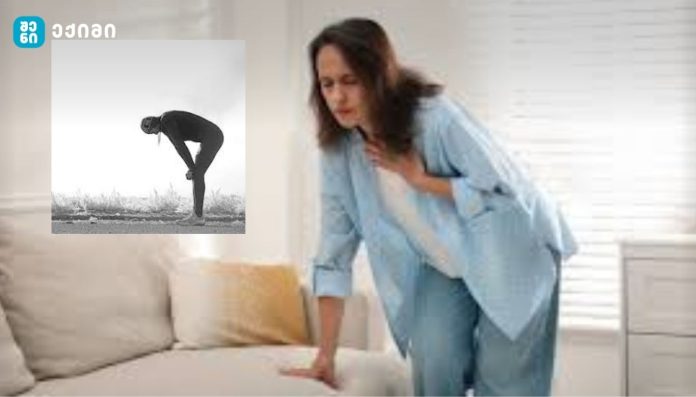 Woman in a blue robe holding her chest while standing beside a sofa, signaling chest pain or discomfort in a home setting.