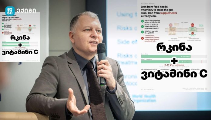 Man in a gray suit speaks into a handheld microphone at a podium during a presentation, with infographic posters visible on both sides in the background.