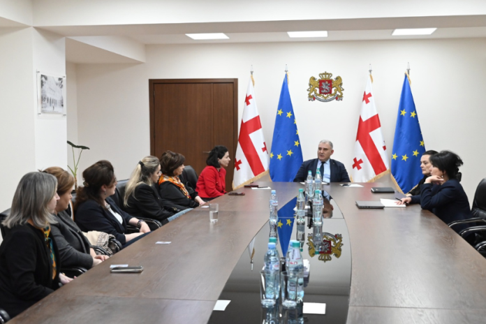 Officials seated around a long conference table in a room with Georgian and EU flags behind a central figure near the head of the table.