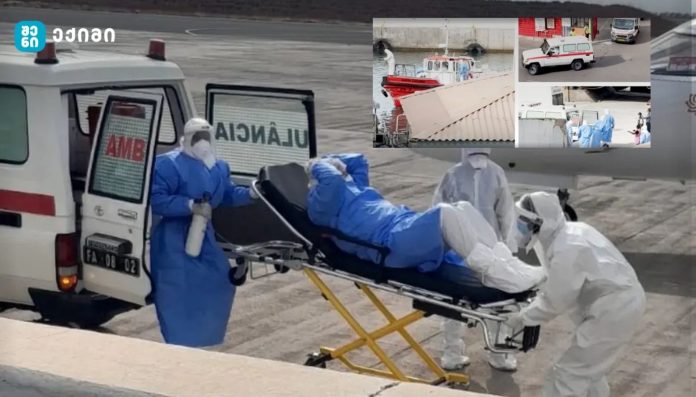 Medical workers in full protective suits transfer a patient on a gurney toward an ambulance on a tarmac site.