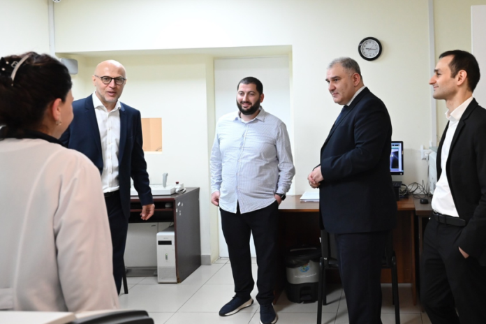 Five professionals, four men in suits and a woman, stand in a bright office discussing near a workstation with a computer and printer in the background.