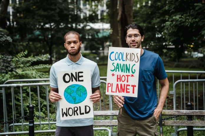 Two young men outdoors hold environmental protest signs; one shows Earth with 'ONE WORLD' text, the other reads 'COOL KIDS SAVING HOT PLANET'.