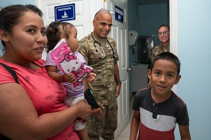 Mother holding a young girl and a boy in a clinic hallway, with two soldiers smiling at the doorway behind them.