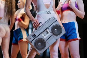 Dancer in a pale pink leotard with a glitter belt carries a large silver boombox among other dancers in colorful athletic wear.
