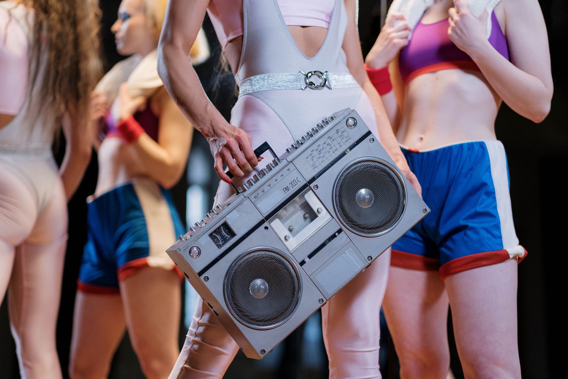 Dancer in a pale pink leotard with a glitter belt carries a large silver boombox among other dancers in colorful athletic wear.