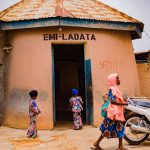Children in colorful outfits stand near an open doorway of a peach building labeled EMI-LADATA; a woman in a pink headscarf passes by with a motorcycle nearby.