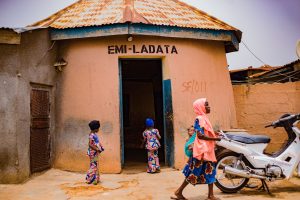 Children in colorful outfits stand near an open doorway of a peach building labeled EMI-LADATA; a woman in a pink headscarf passes by with a motorcycle nearby.