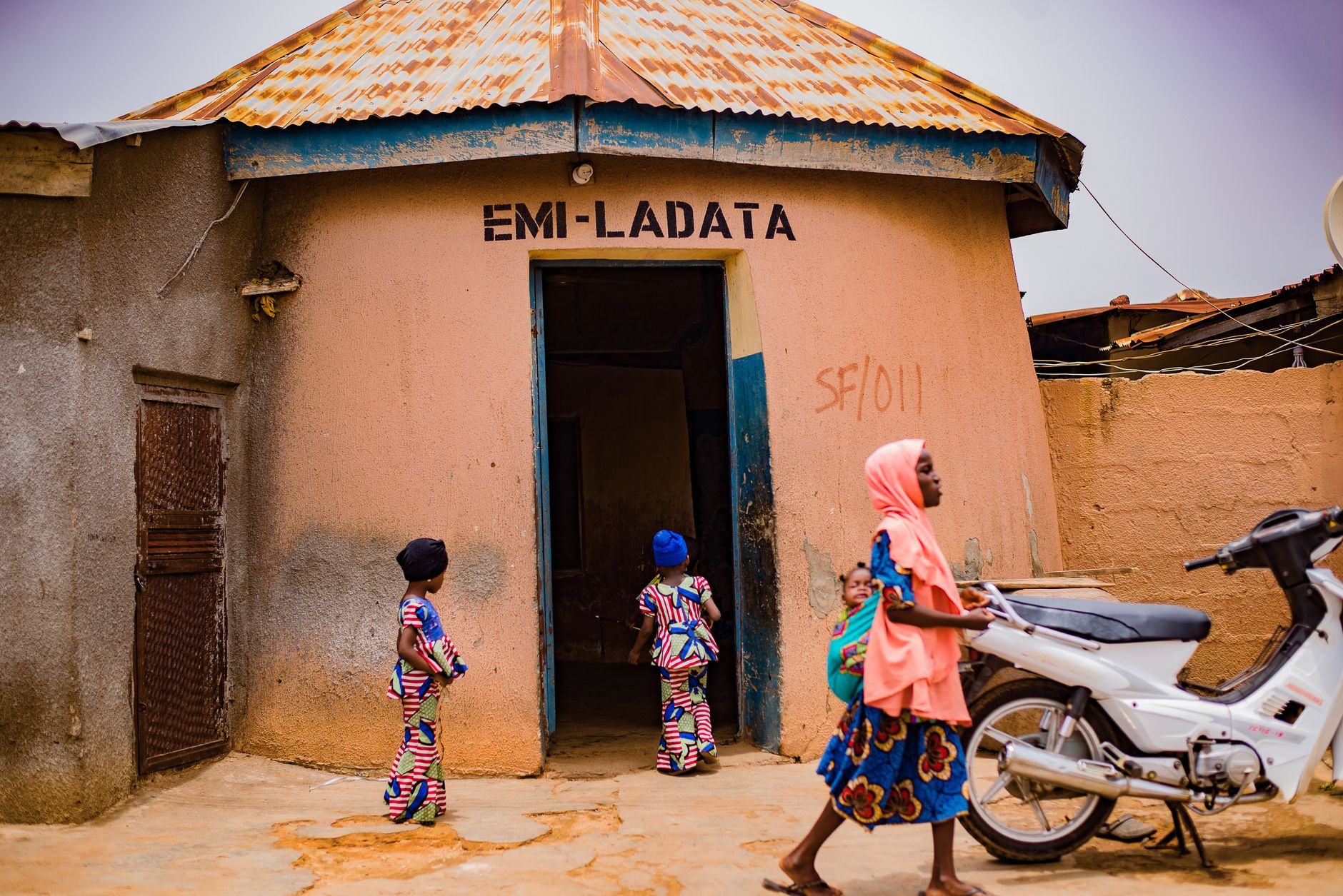 Children in colorful outfits stand near an open doorway of a peach building labeled EMI-LADATA; a woman in a pink headscarf passes by with a motorcycle nearby.