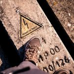 Foot wearing a hiking shoe stepping on a weathered concrete surface with a yellow warning triangle that reads 'Perigo de morte' (Danger of death).