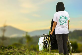 Person in a wheelchair seen from behind, wearing a white t-shirt with a green eco logo, outdoors at sunset.