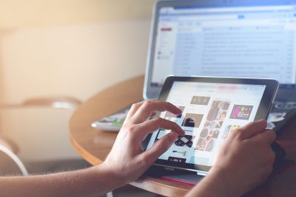 Person browsing an image gallery on a tablet, with a laptop blurred in the background on a wooden table.