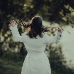Back view of a person in a white outfit outdoors, hair blowing as they flip their head, with greenery in the blurred background.