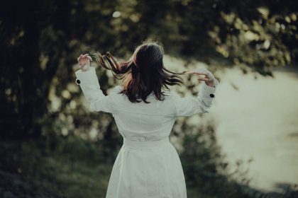 Back view of a person in a white outfit outdoors, hair blowing as they flip their head, with greenery in the blurred background.
