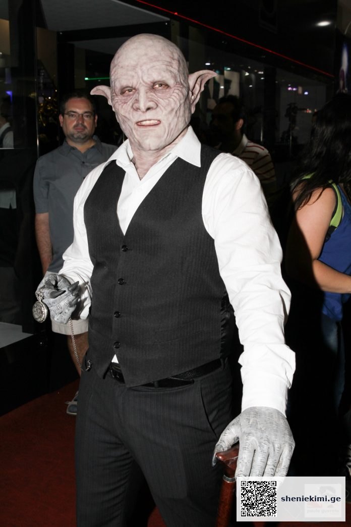 Person wearing a pale goblin mask with pointed ears, a white shirt, dark vest, gloves, and holding a cane.